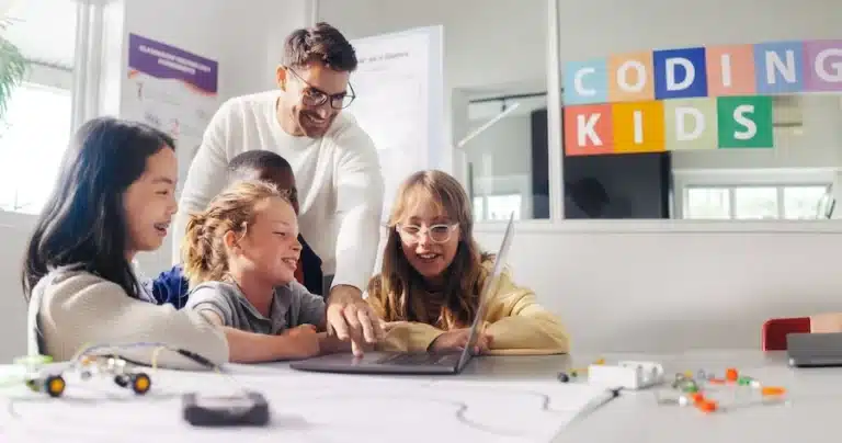 An image showing children with a teacher learning how to code. A sign in the background reads coding kids.