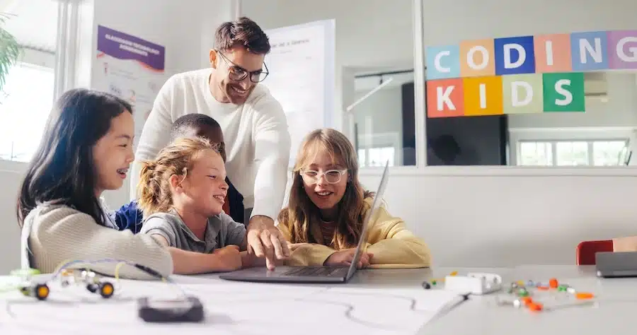 An image showing children with a teacher learning how to code. A sign in the background reads coding kids.