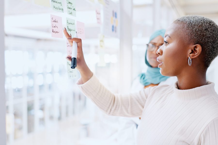 Teamwork, business people and leadership of black woman with sticky note in office workplace. Coaching, collaboration and female employees brainstorming sales or marketing strategy on glass wall