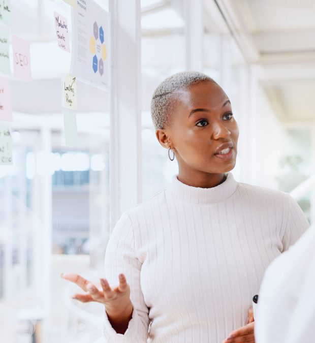 Teamwork, business people and leadership of black woman with sticky note in office workplace. Coaching, collaboration and female employees brainstorming sales or marketing strategy on glass wall