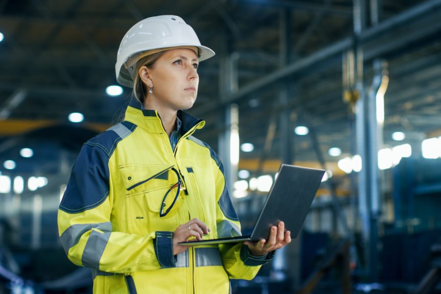 Female Industrial Engineer in the Hard Hat Uses Laptop Computer while Standing in the Heavy Industry Manufacturing Factory. In the Background Various Metalwork Project Parts Lying