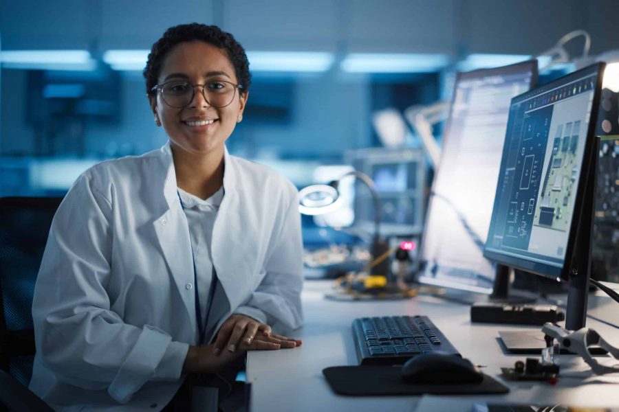 Beautiful Black Latin Woman Wearing Glasses Smiling Charmingly Looking at Camera. Young Intelligent Female Scientist Working in Laboratory. Technological Laboratory in Bokeh Blue as Background